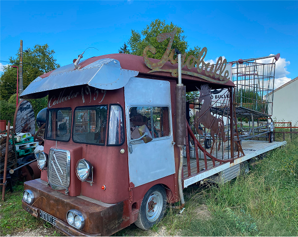 Soon Architecture - Un vieux camion rouge transformé en un char de parade fantaisiste avec un grand panneau en métal indiquant « Isabelle » sur le dessus, des accents métalliques et des sculptures d'animaux sur une plate-forme ouverte derrière la cabine, garé sur l'herbe près des arbres et des bâtiments.