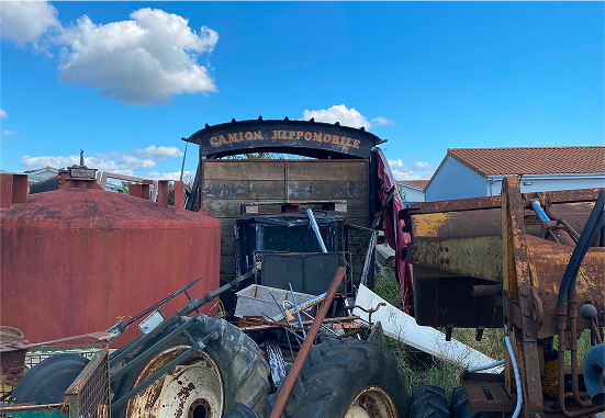 Soon Architecture - Un camion rouillé « Camion Hippomobile » se trouve parmi de vieilles machines, de gros pneus et des débris éparpillés sous un ciel bleu vif avec des nuages, à côté d'un char rouge et d'un bâtiment au toit rougeâtre.