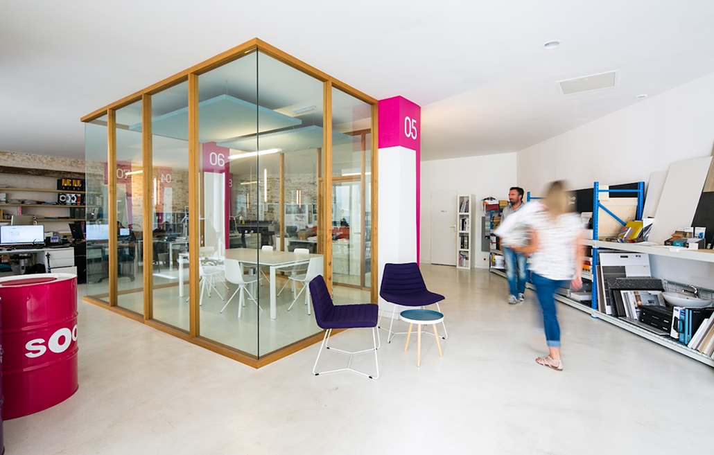 Soon Architecture - Espace de bureaux moderne conçu par l'agence d'architecture Soon, comprenant une salle de réunion vitrée, deux chaises violettes, des touches de blanc et de rose, des étagères remplies de matériaux et deux personnes marchant. Des espaces de travail professionnels et des bureaux sont visibles en arrière-plan.