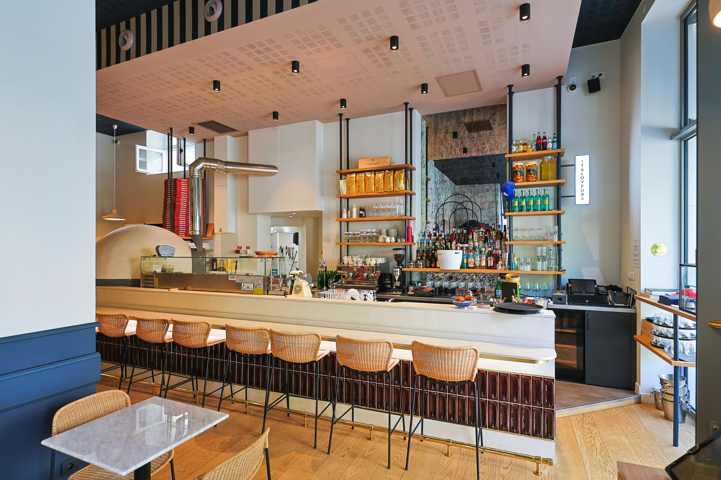 Soon Architecture - Intérieur de café moderne avec un long comptoir, des tabourets en osier, des étagères à bouteilles, des suspensions, un four à pizza et du parquet. De grandes fenêtres laissent entrer la lumière naturelle et une petite table en marbre trône au premier plan.