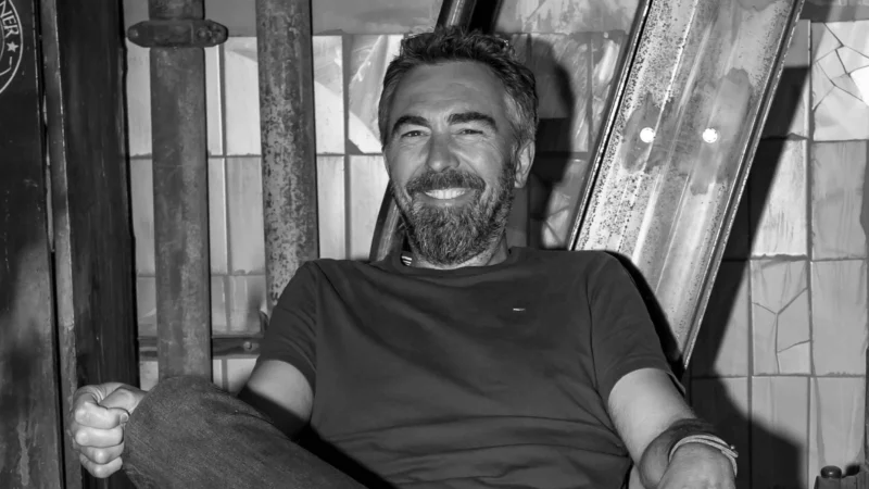 Soon Architecture - Un homme barbu est assis, détendu, une jambe croisée, souriant, dans un décor industriel avec des tuyaux métalliques et des murs carrelés. L'image est en noir et blanc.
