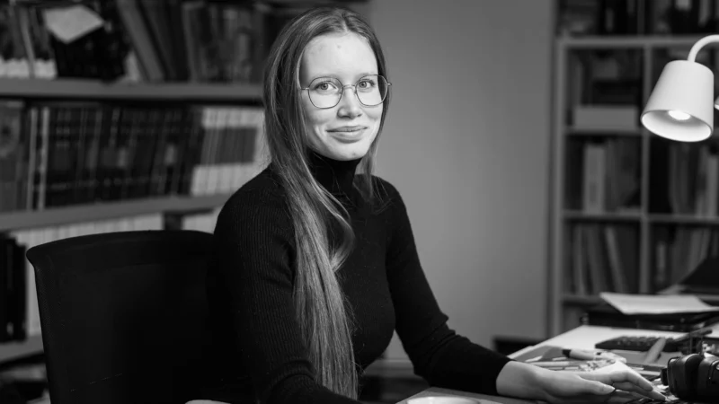 Soon Architecture - Une jeune femme aux cheveux longs et portant des lunettes est assise à un bureau dans un bureau ou une bibliothèque, regardant l'objectif en souriant. Elle porte un pull noir à col roulé et tient une tasse devant elle. Des étagères occupent l'arrière-plan.