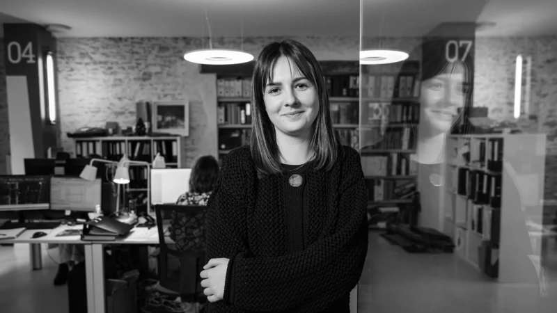 Soon Architecture - Une femme aux cheveux mi-longs sourit dans un bureau, les bras croisés, son reflet sur une paroi vitrée. À l'arrière-plan, des personnes travaillent à des bureaux avec des étagères et des livres derrière. L'image est en noir et blanc.