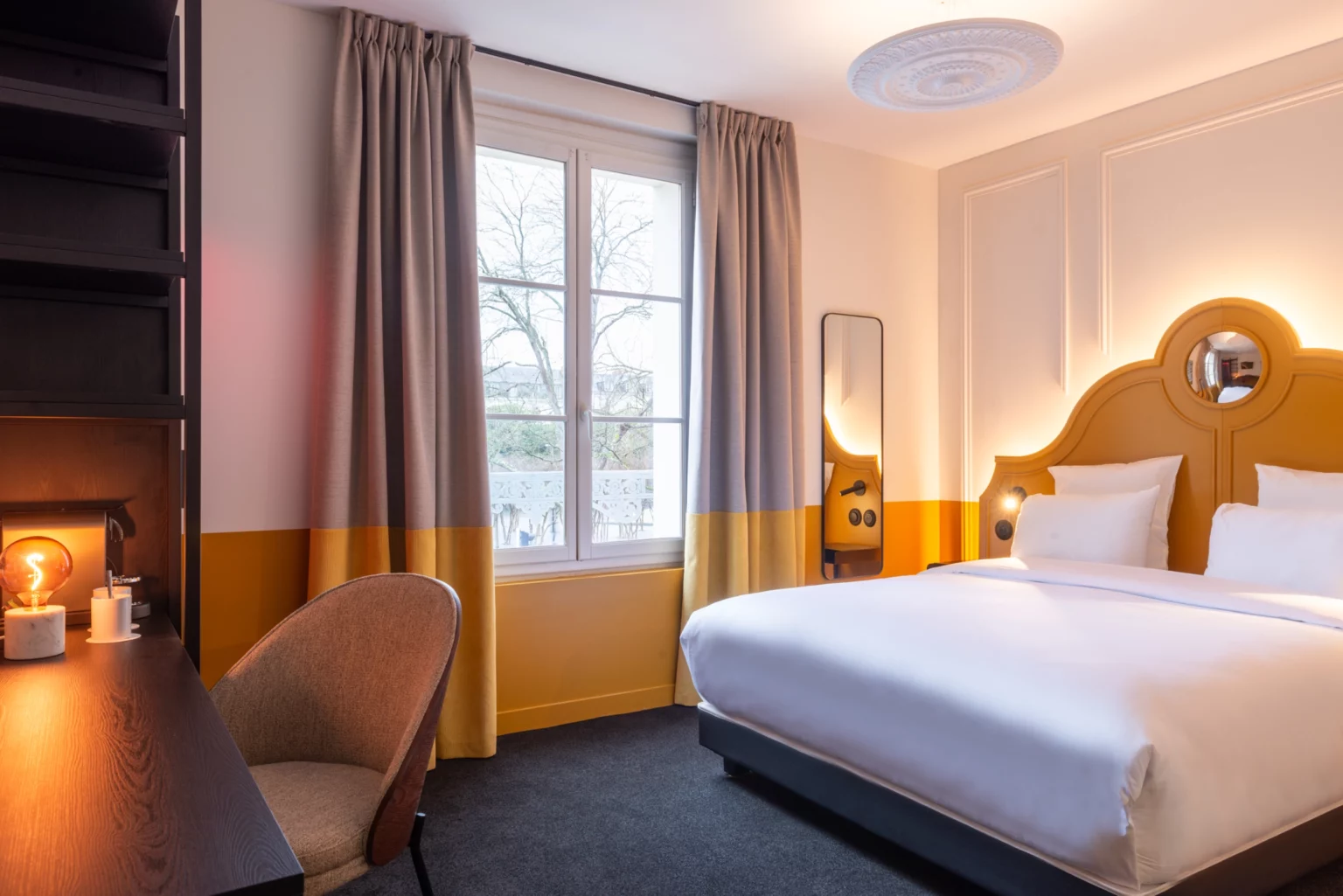 Soon Architecture - Une chambre d'hôtel moderne conçue par Soon architecture comprend un grand lit avec une literie blanche, des murs jaunes et blancs, un bureau professionnel avec une chaise et une lampe, des rideaux jusqu'au sol et une fenêtre laissant entrer la lumière naturelle.