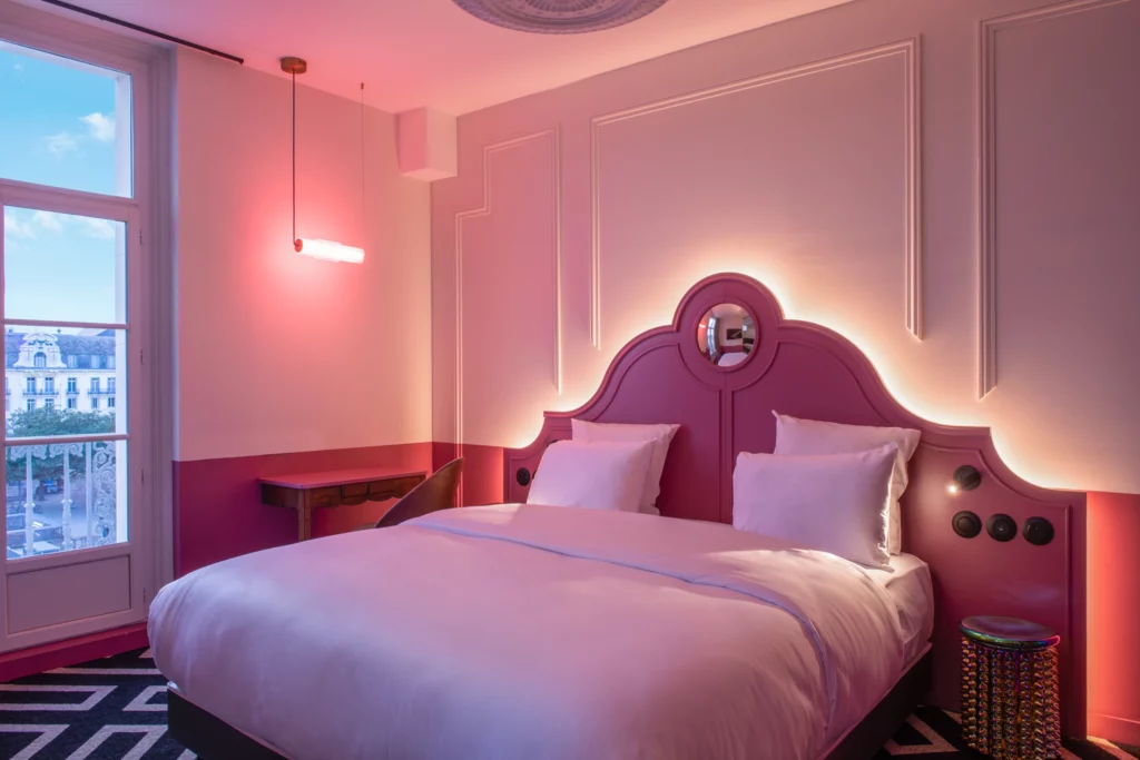 Soon Architecture - Une élégante chambre d'hôtel signée Soon Architecture, avec une tête de lit tapissée de rose, une literie blanche, un éclairage rose doux, un bureau près de la fenêtre et une moquette géométrique noire et blanche. La grande fenêtre offre une vue sur la ville au crépuscule.