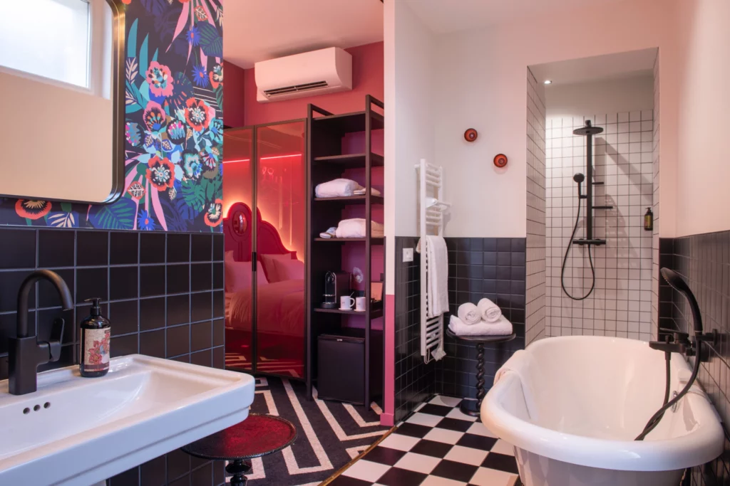 Soon Architecture - Une salle de bains moderne signée Soon Architecture présente un sol à carreaux noirs et blancs, une baignoire îlot, une douche ouverte avec robinetterie noire et des murs aux motifs audacieux. Elle s'ouvre harmonieusement sur une chambre lumineuse aux touches de rouge et aux néons.