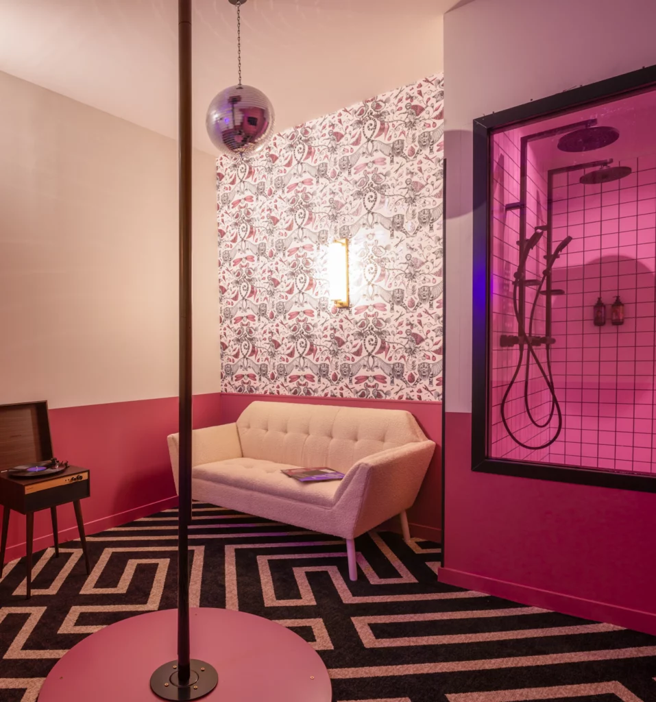 Soon Architecture - Une chambre élégante conçue par Soon architecture comprend un poteau et une boule disco, un canapé blanc, un tapis géométrique noir et blanc audacieux, un tourne-disque sur un support, du papier peint à motifs roses et une douche en verre éclairée par une lumière rose.