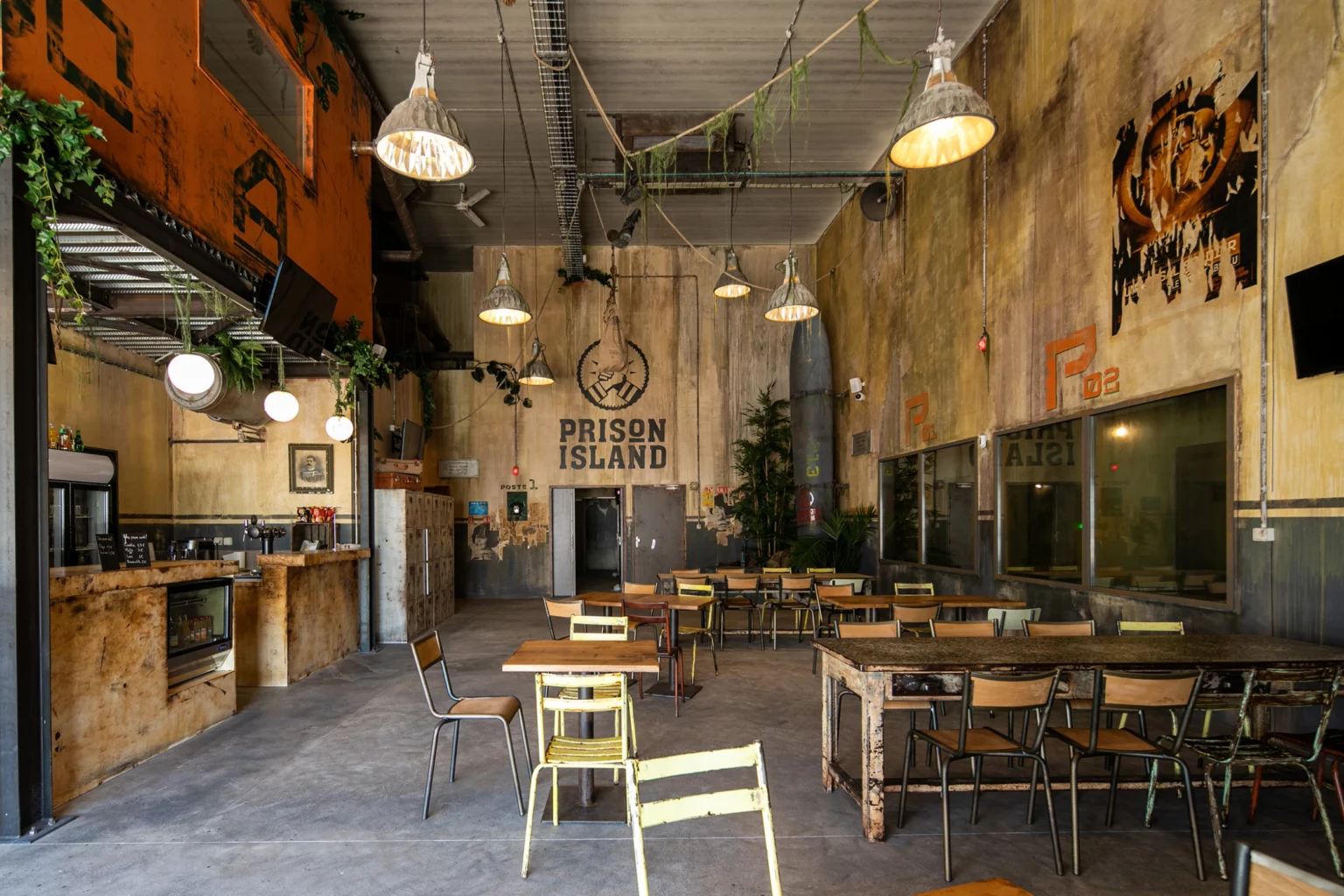 Soon Architecture - Une pièce au style industriel avec des murs patinés, des suspensions, des tables et chaises dépareillées en bois et en métal, des plantes et un grand logo « Prison Island » peint sur le mur du fond. L'espace dégage une atmosphère rustique et urbaine.