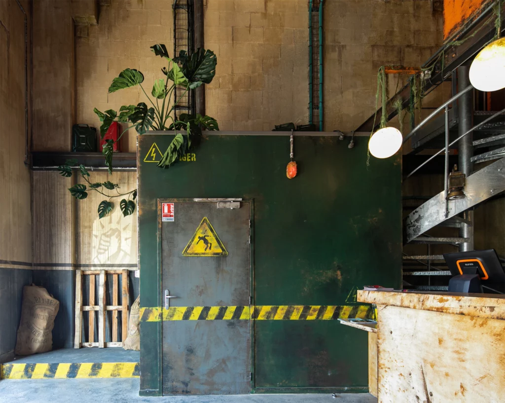 Soon Architecture - Un local industriel verdoyant avec des panneaux d'avertissement, du ruban de précaution, une grande usine et des tuyaux apparents. On aperçoit des escaliers métalliques et des luminaires suspendus, ainsi que des palettes en bois et un sac de sable à proximité.