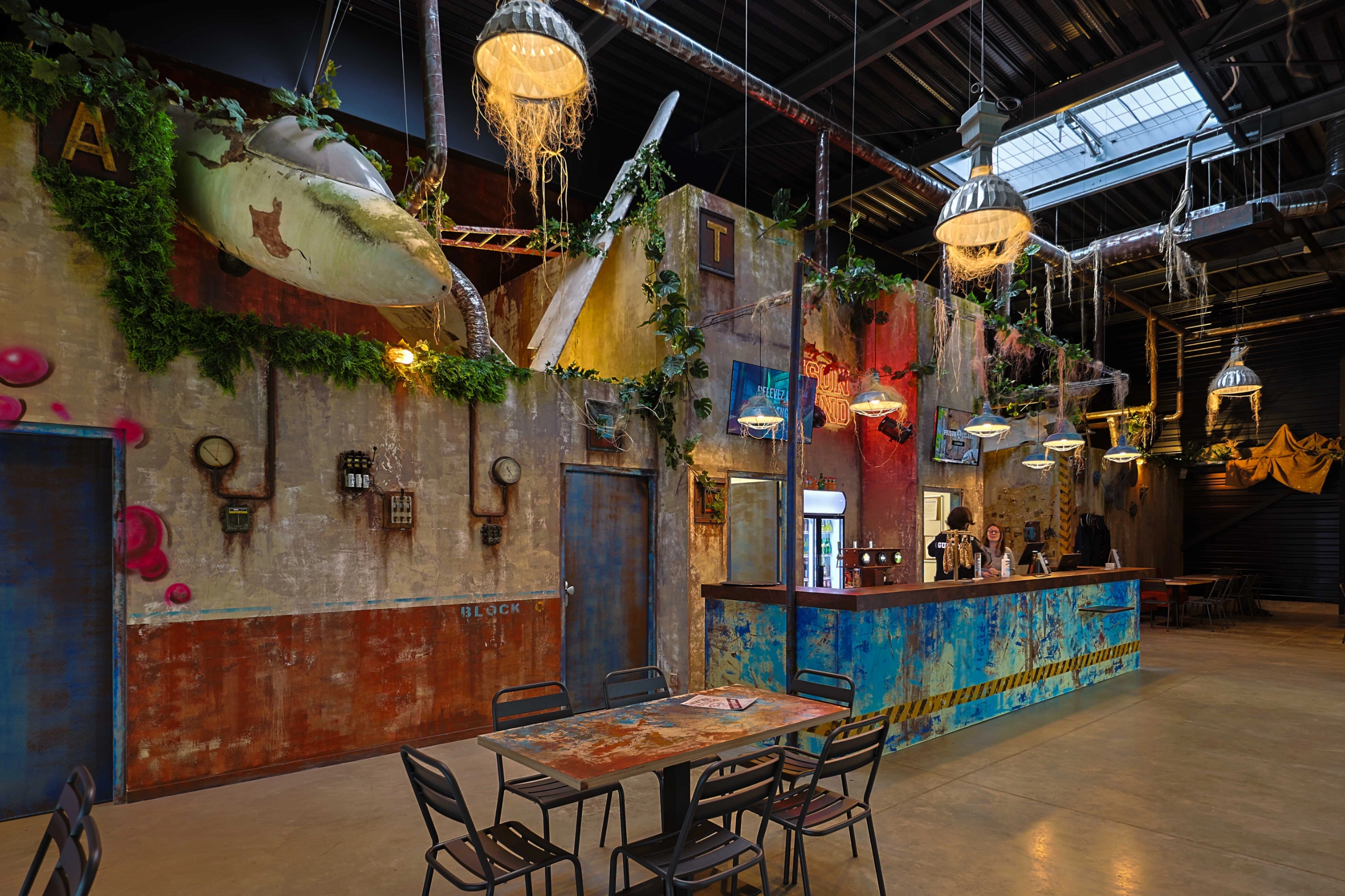 Soon Architecture - Ce bar à thème, décoré dans un style industriel et post-apocalyptique par Soon Architecture, présente des murs usés, des plantes suspendues, de fausses épaves d'avion et des plafonniers originaux. Des tables et des chaises en métal sont disposées près d'un comptoir équipé de robinets.