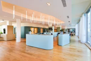 Soon Architecture - Espace de réception de bureau moderne et spacieux avec parquet en bois clair, bureaux incurvés élégants, suspensions suspendues, grandes fenêtres et plusieurs plantes en pot créant une atmosphère lumineuse et accueillante.