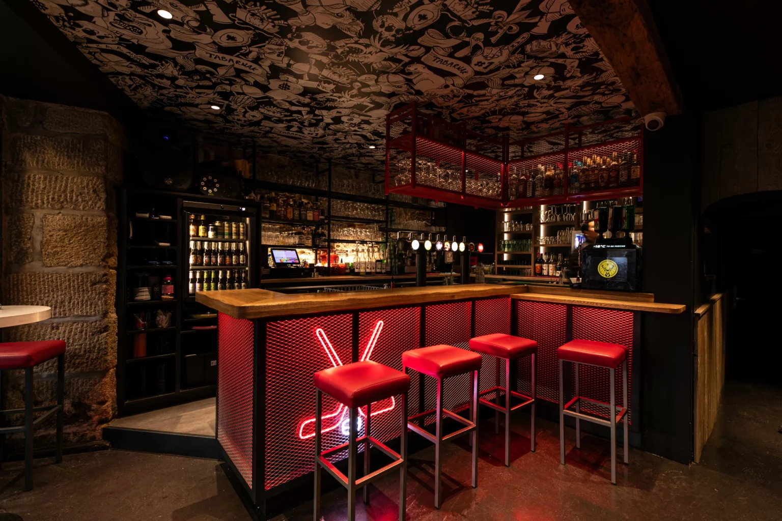 Soon Architecture - Un bar moderne avec un comptoir éclairé en rouge, quatre tabourets rouges, des verres et des bouteilles sur des étagères. Le plafond est orné d'œuvres graphiques en noir et blanc, et les murs sont en pierre et bois foncé. L'atmosphère du bar est élégante et chaleureuse.