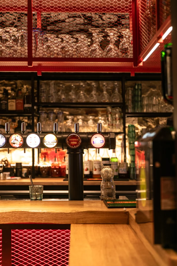 Soon Architecture - Un intérieur de bar avec un comptoir en bois, plusieurs tireuses à bière, des verres suspendus au-dessus et des étagères garnies de bouteilles et de verres en arrière-plan. Le bar est éclairé par une lumière d'ambiance chaleureuse.