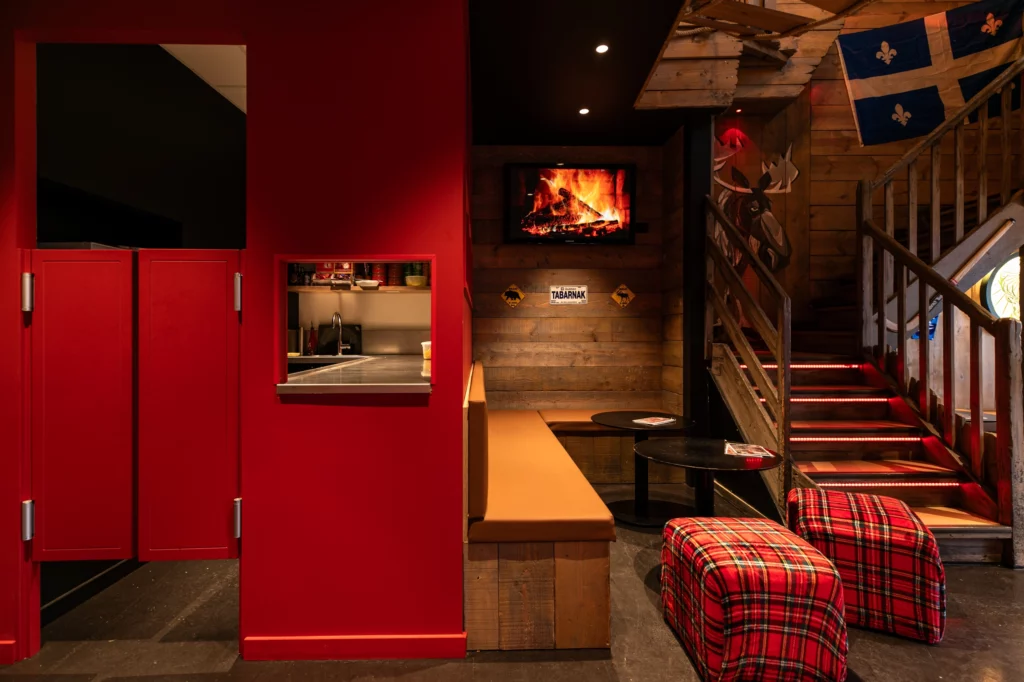 Soon Architecture - Intérieur chaleureux du restaurant avec murs rouges, poufs à carreaux écossais, bancs en bois, petite fenêtre donnant sur la cuisine, escalier et téléviseur diffusant une cheminée. Un éclairage chaleureux met en valeur le décor rustique et chaleureux.