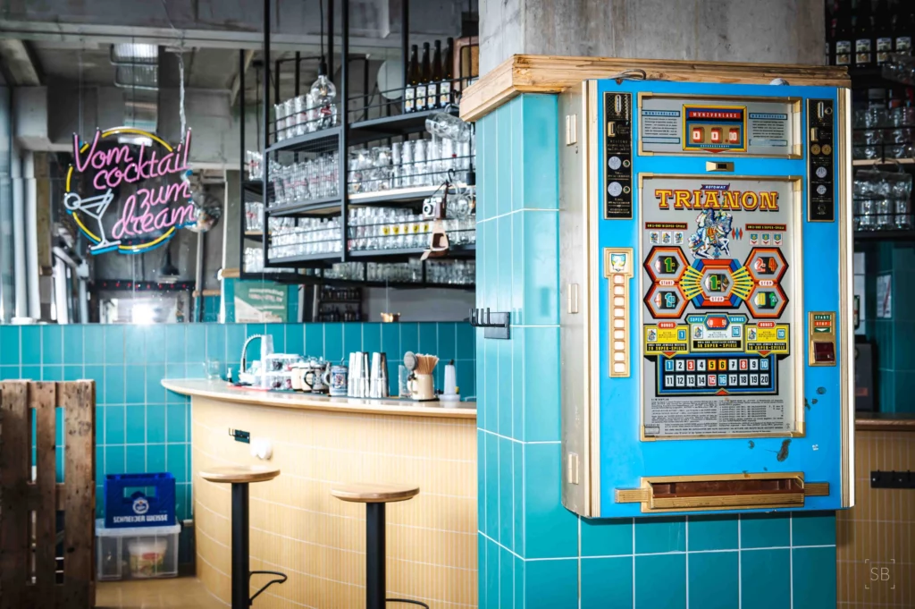 Soon Architecture - Un distributeur automatique vintage aux couleurs vives est installé sur un mur carrelé bleu sarcelle dans un bar élégant conçu par Soon Architecture. En arrière-plan, on aperçoit des verres, des bouteilles et une enseigne lumineuse indiquant « Vom cocktail zum dream ».