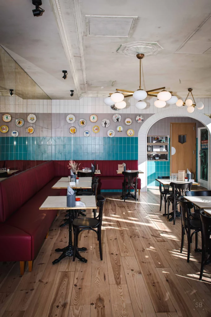 Soon Architecture - Café lumineux de style rétro, conçu par Soon Architecture, avec murs carrelés bleu sarcelle et blanc, plafonniers ronds, parquet, bancs rouges, chaises noires et assiettes décoratives. Les tables sont dressées pour une ambiance à la fois professionnelle et chaleureuse.