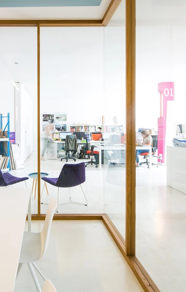 Soon Architecture - L'intérieur de bureau moderne de l'agence d'architecture Soon présente des murs en verre, un sol blanc et des chaises colorées. Des équipes de professionnels travaillent à des bureaux dans un espace ouvert et lumineux, avec des étagères et un panneau rose étiqueté « 01 » en arrière-plan.