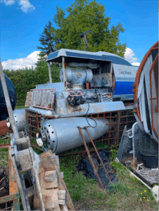 Soon Architecture - Une grande machine patinée par les intempéries, portant l'inscription « Techno », trône dans une cour envahie par la végétation. Elle semble vieille et partiellement démontée, entourée de palettes en bois, de structures métalliques et de hautes herbes.