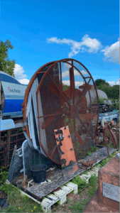 Soon Architecture - Une grande structure de ventilateur en métal rouillé se dresse à l'extérieur sur des palettes en bois, entourée d'équipements divers et de végétation sous un ciel bleu vif avec quelques nuages.
