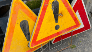 Soon Architecture - Trois panneaux d'avertissement triangulaires usés, ornés d'un point d'exclamation au centre, sont empilés sur une surface rugueuse en gravier, à l'extérieur. Jaunes avec des bordures orange ou rouges, ils présentent des traces de saleté et de rouille.