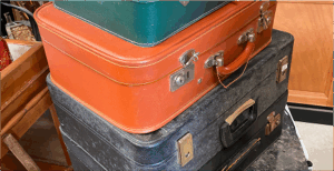 Soon Architecture - Une pile de trois valises vintage : une valise verte sur le dessus, une orange au milieu avec une poignée en cuir et une plus grande valise noire usée en dessous, le tout reposant sur une surface en marbre.