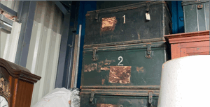 Soon Architecture - Deux grandes malles vertes usées, numérotées 1 et 2, sont empilées dans un garde-meuble. Un cadenas est accroché à la malle supérieure. Divers objets, dont une horloge et une lampe, entourent les malles.