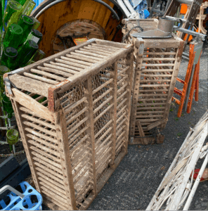 Soon Architecture - Deux caisses en bois patinées avec des côtés à lattes et du grillage reposent sur l'asphalte parmi divers objets, notamment des bouteilles vertes empilées, des tiges métalliques et des chaises orange, suggérant un décor de marché aux puces ou de vente de garage.