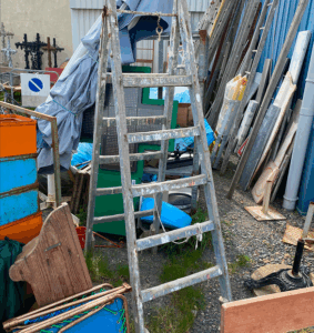 Soon Architecture - Une échelle en métal patiné se dresse parmi des matériaux de construction variés, des planches de bois, du tissu et de l'équipement dans une zone de stockage extérieure.