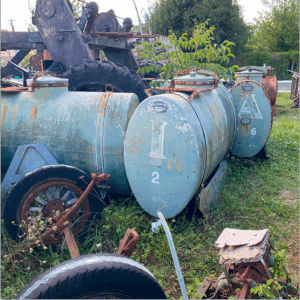 Soon Architecture - Deux vieux réservoirs métalliques rouillés, étiquetés I 2 et A 6, sont posés dans l'herbe envahie par la végétation, à proximité de roues et de pièces de machines mises au rebut, avec un tracteur et des arbres en arrière-plan.