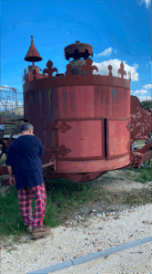 Soon Architecture - Une personne vêtue d'une chemise noire et d'un pantalon à carreaux rouges se tient à côté d'une grande structure métallique ronde et rouillée avec des bords décoratifs, à l'extérieur sur une surface de gravier sous un ciel bleu.